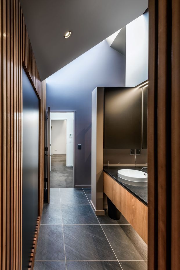 A contemporary bathroom vanity viewed from a narrow hallway or entry passage. The space features dark large-format floor tiles, a timber slatted wall and matching timber vanity cabinetry, a dark stone-like benchtop, white vessel basin, large dark mirror, and deep blue-grey walls. A doorway opens to an adjoining carpeted room, while concealed and recessed lighting creates a refined, moody atmosphere.