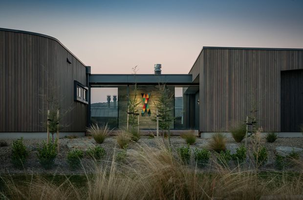 Single-storey contemporary building exterior with dark vertical timber cladding, strong rectilinear wings, a glazed central entry or connector, and a low flat roof. The building is set within a drought-tolerant landscaped garden featuring ornamental grasses and sparse planting, photographed in soft evening light.