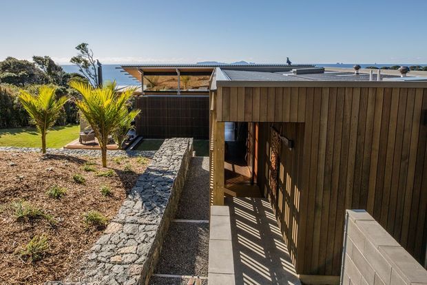 Exterior view of a low-profile contemporary coastal house, with dark vertical timber cladding, a flat metal roof, sheltered outdoor circulation, and textured stone retaining walls. The landscaped garden and ocean horizon are visible beyond the building.
