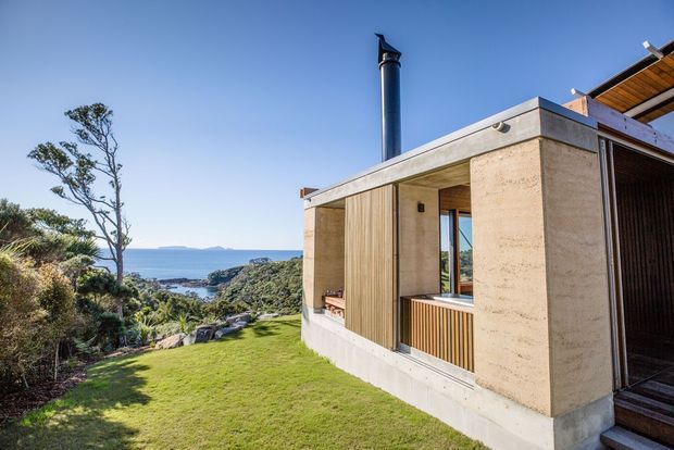 Exterior view of a contemporary coastal house set into a landscaped hillside, with a flat concrete roof, pale textured concrete walls, vertical timber screening, deep recessed openings, and a prominent dark metal chimney. The building overlooks dense coastal vegetation and the sea beyond, with a bright blue sky and lawn in the foreground.