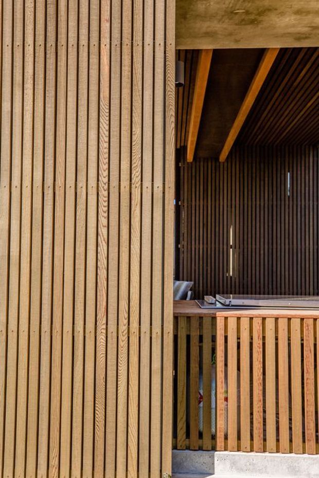 Close-up architectural view of a timber-clad exterior and covered outdoor deck or balcony. The facade uses closely spaced vertical timber battens, continued across the ceiling and balustrade, with exposed timber beams and a recessed seating area.