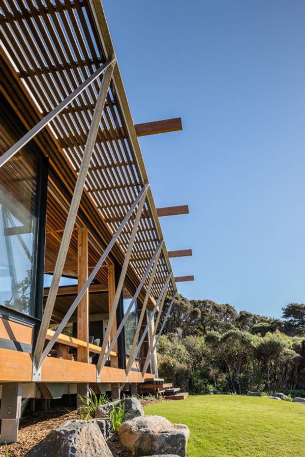 Exterior architectural photograph of a contemporary timber-clad building set within a landscaped bush setting. The facade features extensive vertical timber slatting forming a screened upper structure, exposed timber posts and beams, large glazing, diagonal bracing, projecting timber members, and a raised deck or platform. The building sits beside a manicured lawn and native-looking vegetation under a clear blue sky.