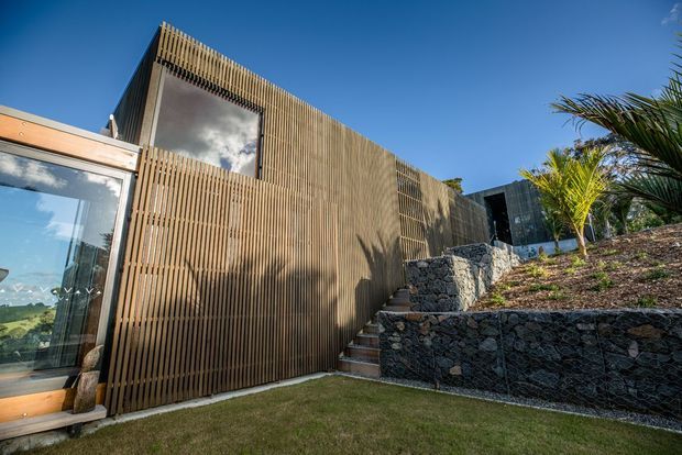 Exterior view of a contemporary, low-profile building on a landscaped sloping site. The facade is predominantly clad in closely spaced vertical timber battens, with large glazed openings and a timber-framed glazed section at left. Stone-filled gabion retaining walls and a stairway connect different garden levels, with tropical planting visible at right under a clear blue sky.