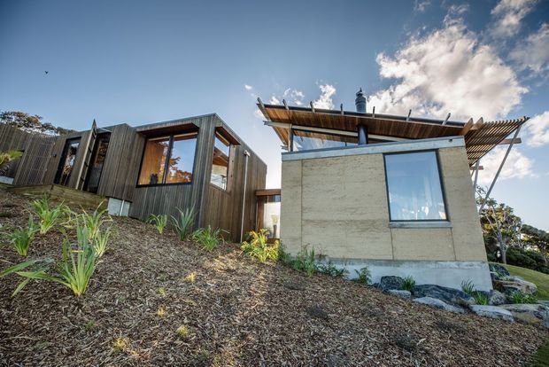 Exterior view of a modern, compact residential building on a sloped landscaped site. The structure combines dark vertical timber cladding, exposed concrete walls, large windows, and a projecting timber-framed roof canopy with a chimney. Native-style planting and mulch surround the building.