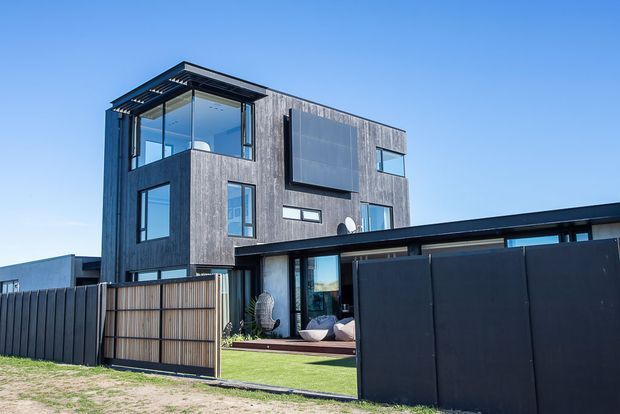 Exterior view of a contemporary multi-storey residence with dark vertical timber cladding, extensive black-framed glazing, projecting upper-level volumes and a flat-roofed lower pavilion. A tall dark boundary fence, timber gate and small landscaped outdoor area frame the foreground.