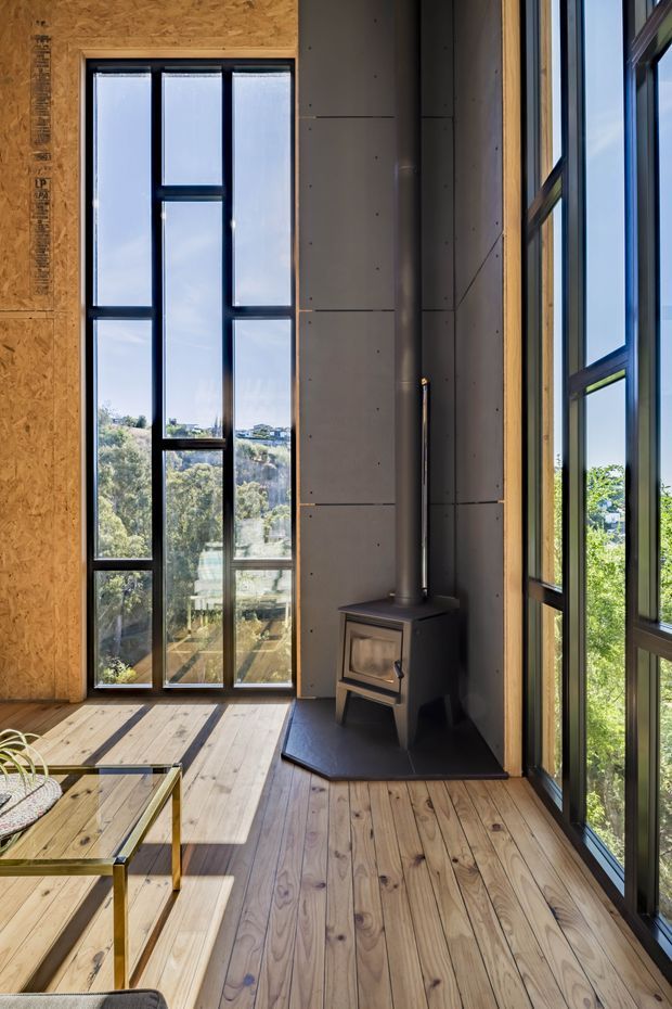 A tall, light-filled interior corner with full-height black-framed glazing overlooking a leafy landscape. The room features wide timber flooring, exposed OSB wall sheathing, dark concrete wall panels, a compact freestanding wood-burning stove with flue, and a simple glass-topped coffee table. The vertical glazing and warm timber create a contemporary, natural atmosphere.