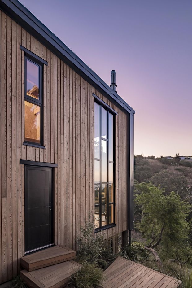 Exterior view of a compact, vertically clad timber building on a steep, vegetated site. The facade has dark metal window and door frames, tall glazing, a pitched roof edge with a flue, and a timber deck and entry steps overlooking the surrounding landscape at dusk.