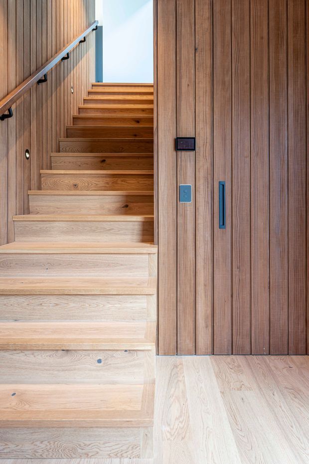 A bright, contemporary stairway lined with extensive warm timber detailing. Broad pale timber treads rise beside a darker vertical timber-paneled wall and concealed storage door, with a simple timber handrail and black metal brackets. A small wall control panel and narrow dark door pull provide subtle modern accents.