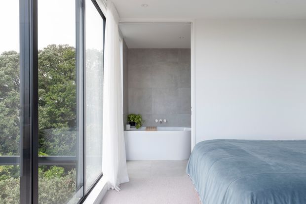 Bright contemporary bedroom with a large floor-to-ceiling window wall overlooking trees, sheer white curtains, pale carpet, and a partially enclosed white freestanding bathtub set against a large-format grey tiled wall. The bed occupies the foreground with soft blue-grey bedding, creating an airy, calm atmosphere.