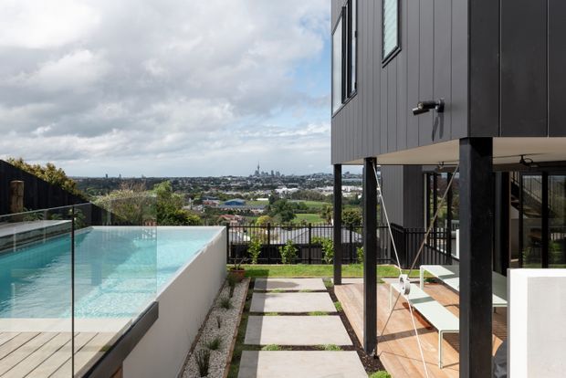 A contemporary elevated residential exterior overlooking a suburban cityscape. The image shows a turquoise swimming pool with frameless glass balustrades on the left, a paved stepping-stone walkway through a narrow landscaped strip, and a dark-clad multi-level house with large glazed openings, slender black structural posts, and a covered outdoor terrace on the right. The distant urban skyline and hilly neighbourhood are visible beyond the property.