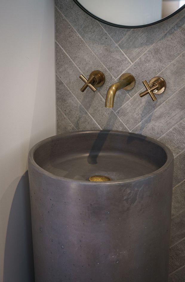 Close-up of a sculptural bathroom vanity featuring a large cylindrical concrete vessel basin, wall-mounted brushed brass cross-handle tapware, and a grey textured tile splashback laid in a diagonal pattern. A round mirror is partially visible above.