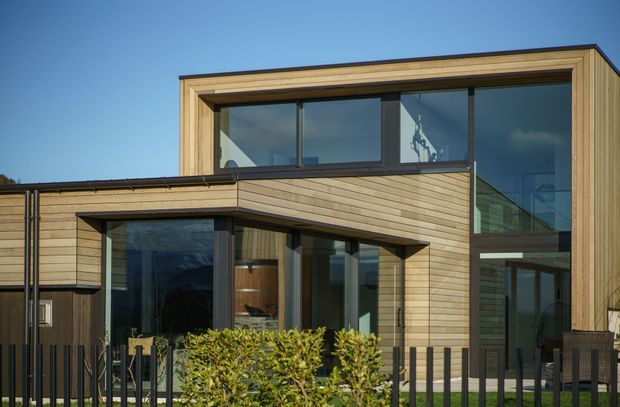 Exterior view of a contemporary two-storey home with clean rectilinear volumes, extensive dark-framed glazing, and horizontal light timber cladding. A projecting lower-level section creates a sheltered outdoor area, with a small landscaped frontage and black metal fencing.