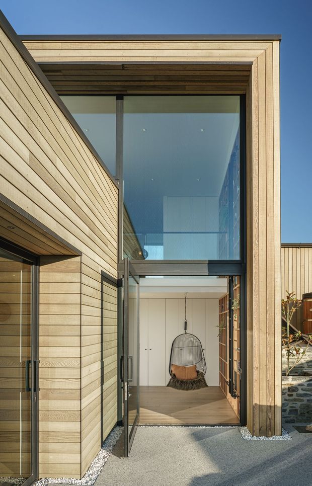 A modern timber-clad residential entry courtyard with a dramatic double-height glazed opening. Pale horizontal timber boards frame the facade and soffit, while dark metal window and door frames contrast with the warm wood. Through the open glass entry, a compact interior foyer with pale cabinetry, timber flooring, and a hanging chair is visible. Gravel edging, concrete paving, and a low stone wall complete the exterior approach.