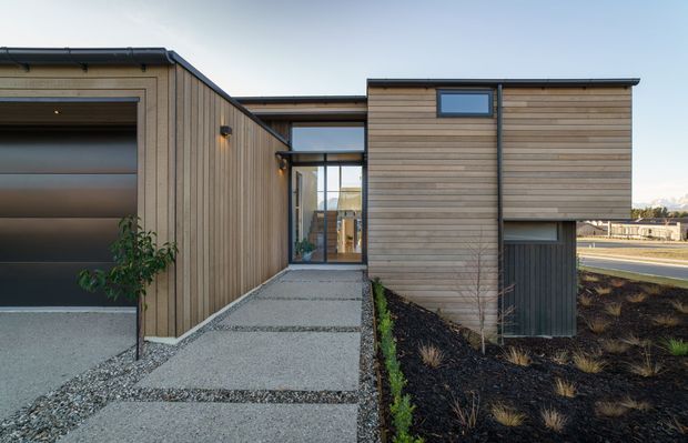 Contemporary single-storey building exterior with horizontal and vertical timber cladding, dark metal roof edges and trims, and a glazed entry connection. A textured concrete path leads between the timber-clad volumes, with a planted garden bed and roadway visible beyond.