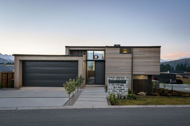 Front exterior view of a contemporary multi-level residence with a prominent attached single-car garage, horizontal timber-clad upper facade, dark metal-framed glazing, and a stone-clad entry feature wall. The property sits on a suburban street with mountains visible in the distance.