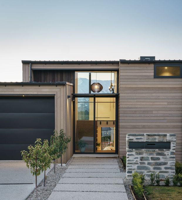 Front exterior of a contemporary single-storey residence with horizontal timber cladding, a dark sectional garage door, tall glazed entry, flat rooflines, and a masonry feature wall beside a concrete path. The facade has a restrained neutral palette and strong vertical entry glazing.
