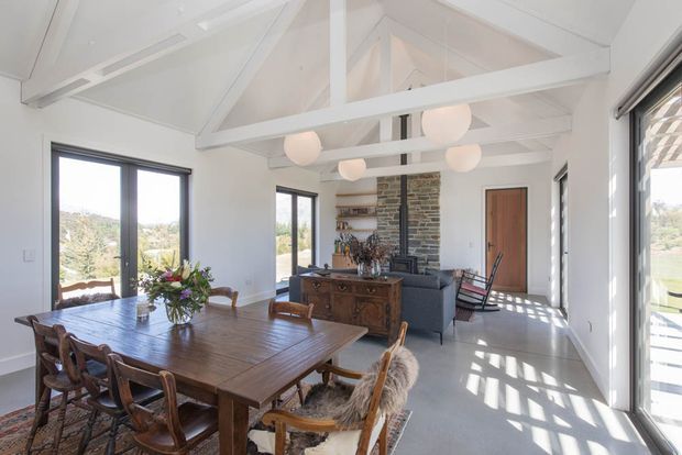 Bright open-plan dining and living area with a large rustic timber dining table and chairs in the foreground, a seating area beyond, and a stone-clad fireplace wall with built-in shelving. White pitched ceiling with exposed beams, clerestory glazing, pendant globe lights, timber-framed doors, and extensive windows create an airy, contemporary farmhouse-like interior.