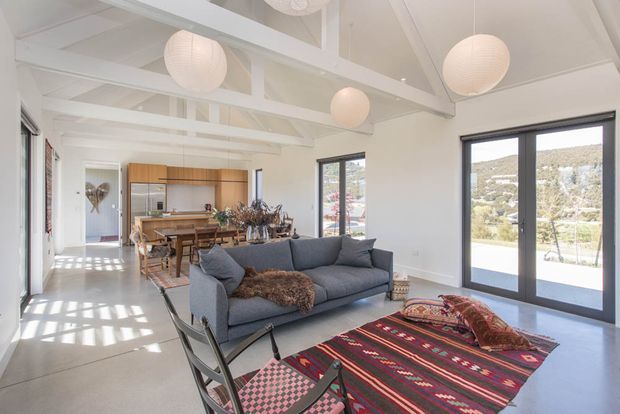 Bright open-plan interior combining a living room and dining/kitchen area. The space has white walls and exposed white-painted ceiling beams, large dark-framed glazed doors and windows, pale hard flooring, a charcoal sofa, timber dining furniture, and colourful patterned rugs. Multiple spherical pendant lights hang from the high, beamed ceiling, with a sunlit hillside visible outside.