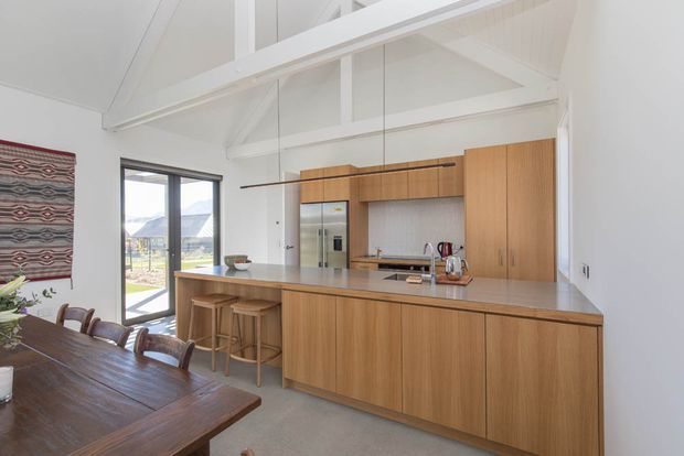 A bright open-plan kitchen and dining area with a large timber island, integrated cabinetry, stone benchtop, bar stools, and a timber dining table. The kitchen has a high pitched white ceiling with exposed structural beams, pendant lighting, stainless-steel appliances, and large glazing opening toward a mountainous landscape. The space feels warm, airy, and contemporary with natural timber joinery and neutral finishes.