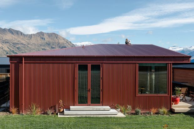 Single-storey compact dwelling with a dark red corrugated metal-clad facade and low-pitched red metal roof. Full-height ribbed vertical cladding, glazed double doors, and a wide window face a small lawn and planted entry. Mountain ranges are visible behind the building, with neighbouring structures at either side.