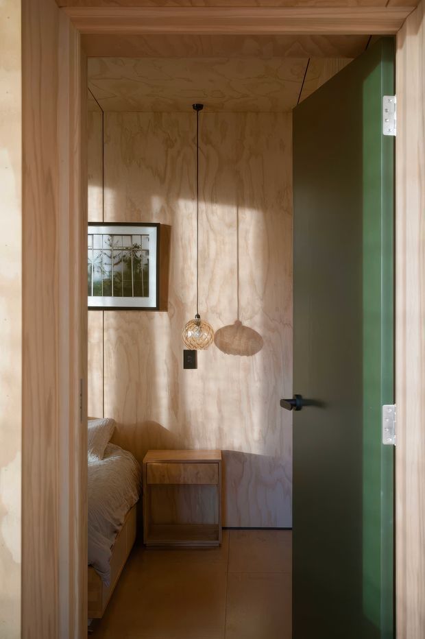 View through a timber-framed doorway into a compact bedroom clad in warm plywood wall and ceiling panels. The room contains a partially visible bed, a small timber bedside table, a framed artwork, and a pendant light with a woven spherical shade. A deep green door with dark hardware stands open, creating contrast against the natural timber interior.