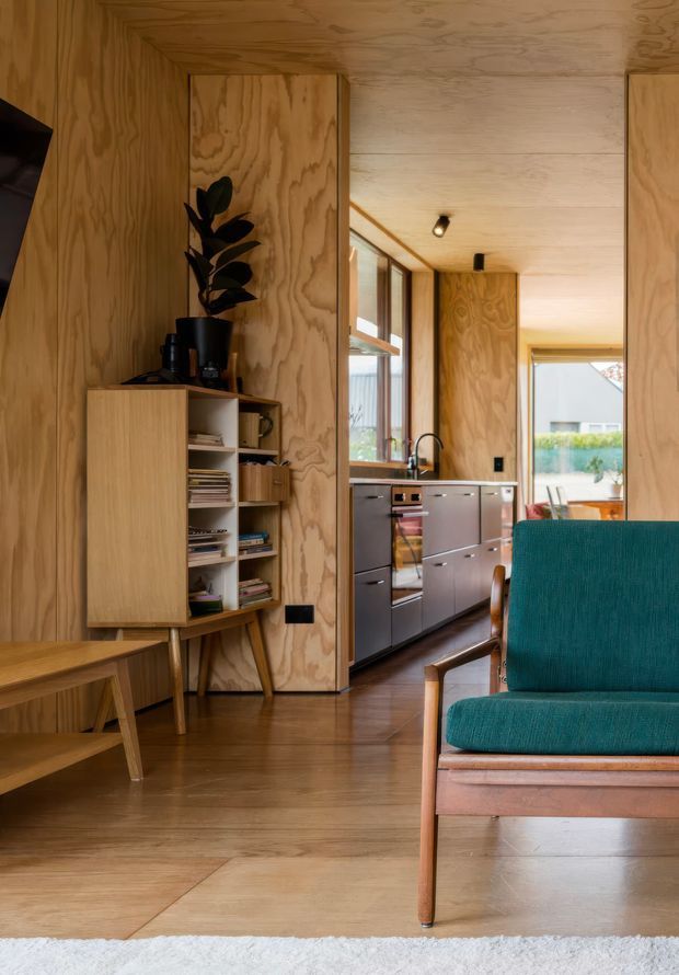 Interior view of a compact, warm timber-lined home showing a mid-century-style teal upholstered armchair in the foreground, a timber dining table and open shelving, and a galley kitchen beyond. Plywood wall, ceiling and cabinetry surfaces create a cohesive natural-wood interior, with dark kitchen cabinetry, stainless appliances, timber flooring, and generous windows bringing in daylight.