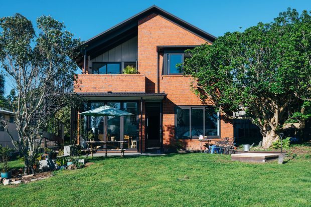 A two-storey residential house viewed from the rear garden, featuring exposed red brick walls, dark metal-framed windows and doors, a steep gabled roof, and a covered outdoor patio with an umbrella. Mature trees and a lawn surround the home.