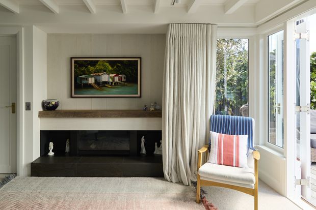 Bright, refined living room with a built-in fireplace wall, pale panelled cabinetry or wall lining, exposed white ceiling beams, and a large set of windows and glazed French doors. Full-height cream curtains frame a blue-and-coral upholstered armchair, while a neutral rug, timber-toned mantel, dark hearth, and framed artwork complete the room.