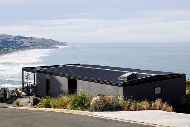 Exterior view of a contemporary single-storey home on a coastal, sloping site. The dark low-profile building has a flat roof, corrugated-looking dark cladding, extensive corner glazing, and a roof skylight. Native-style grasses and shrubs landscape the frontage, with an expansive ocean and rugged coastline visible beyond.