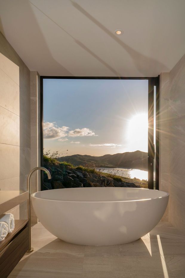 A refined bathroom with a freestanding white oval bathtub positioned in front of large black-framed glazing. Pale stone or porcelain tiles cover the walls and floor, with a brushed metal floor-mounted bath filler and recessed ceiling light. The opening frames a dramatic sunlit mountain and lakeside landscape.