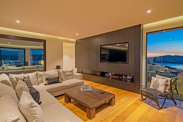 Warm, contemporary living room with large cream sectional sofas, a substantial timber coffee table, timber flooring, and a dark timber feature media wall incorporating a television and linear fireplace. Recessed ceiling lighting and concealed cove lighting create a cozy atmosphere, while expansive glazing provides a view toward water and hills. A woven lounge chair sits beside the window.
