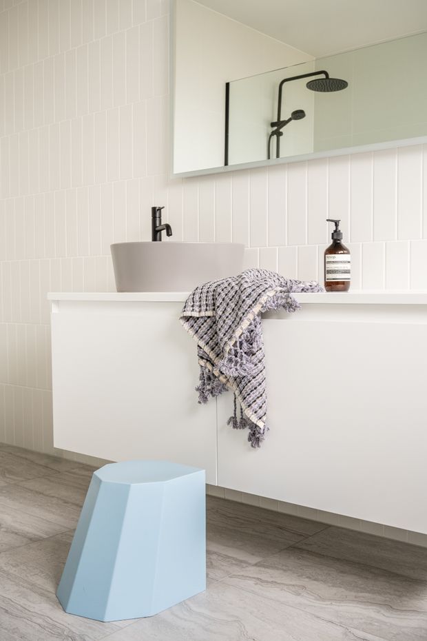 A bright, contemporary bathroom featuring a wall-mounted white vanity with a vessel basin, matte black tapware, a large frameless mirror, and a walk-in shower reflected in the mirror. White vertical wall tiles are paired with grey stone-look floor tiles. A pale blue geometric stool and a textured monochrome towel add subtle accents.