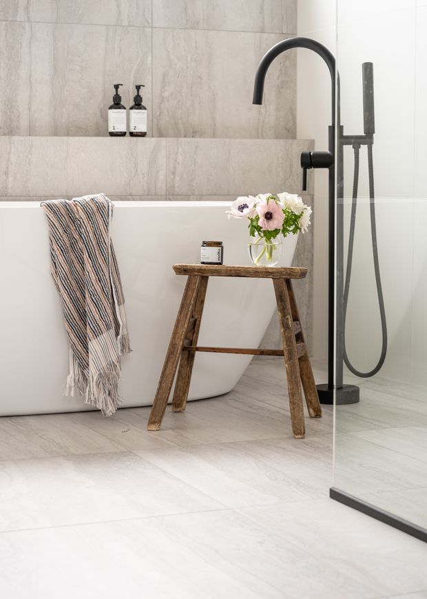 A bright, refined contemporary bathroom featuring a freestanding white bathtub, large pale stone-look wall and floor tiles, matte black curved tapware and shower fittings, and a clear glass shower screen. A rustic timber stool with flowers and a folded striped towel adds warmth and contrast.