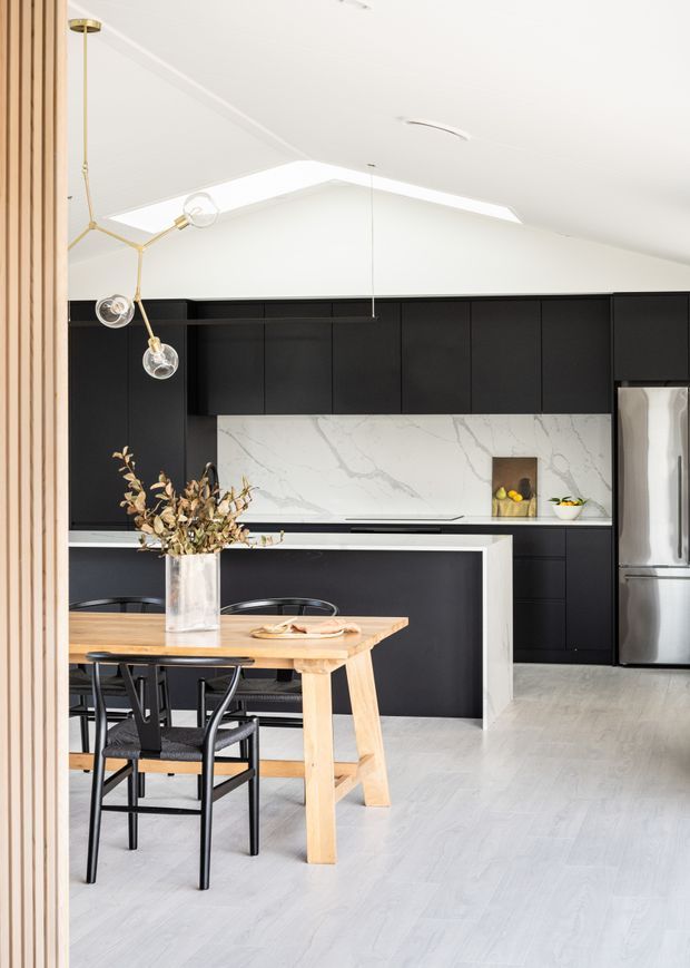 Bright contemporary kitchen and dining area with matte black full-height cabinetry, a white marble-look splashback, central island, pale timber dining table, black dining chairs, and stainless-steel appliances. The room has a pitched ceiling with skylight-like clerestory glazing and a sculptural brass-and-glass pendant light.