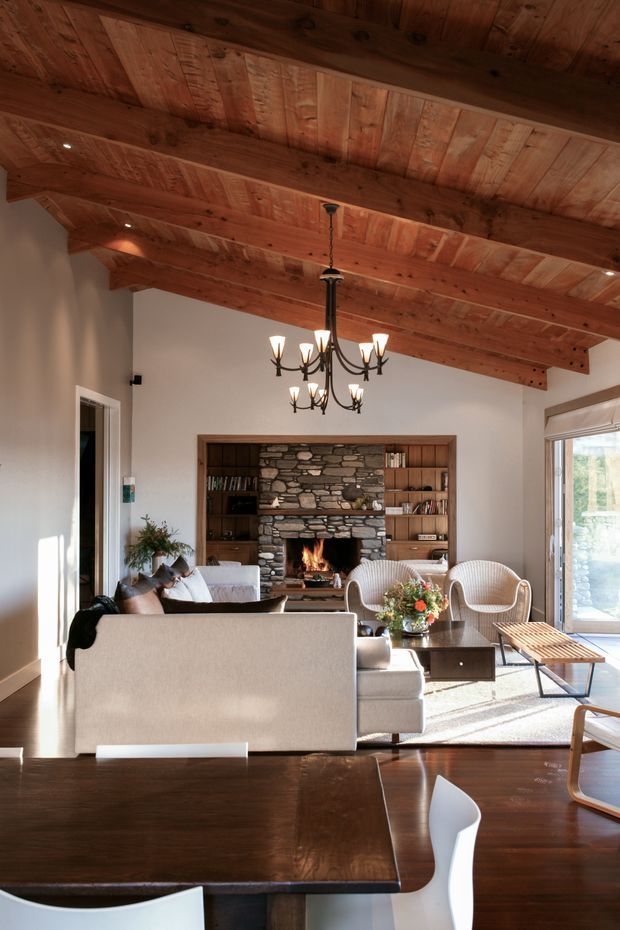 A warm, rustic living room viewed from an adjacent dining area. The room features a dramatic exposed timber-beam and timber-board ceiling, pale plaster walls, a large stone fireplace with built-in dark timber shelving, neutral upholstered sofas and lounge chairs, a dark timber coffee table, and a traditional black multi-arm chandelier. Large glazing at the right brings in natural light and views of greenery.