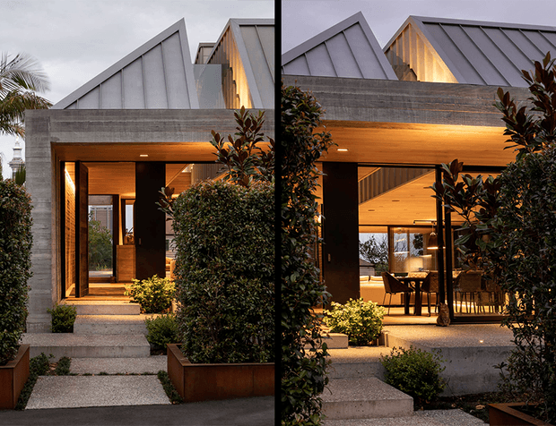 Split-view architectural exterior of a contemporary residence at dusk, showing a textured concrete facade, warm timber-lined entry and covered outdoor living area, landscaped planting, and sharply angled standing-seam metal roof forms. Glazing reveals part of the interior dining/living space.