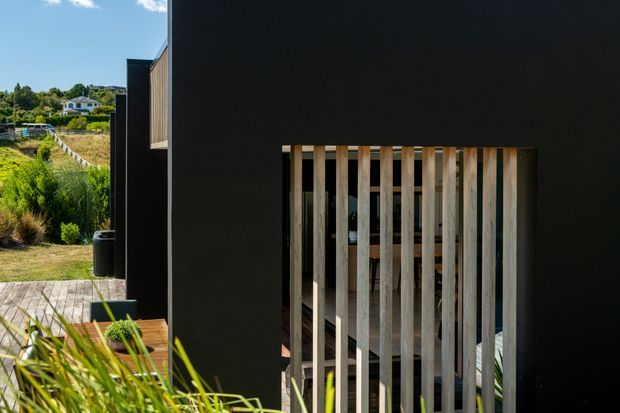 Exterior architectural view of a contemporary dark-clad building, featuring a timber vertical slatted screen over an opening or entry area. A timber deck and landscaped planting are visible beside the building, with a residential hillside setting in the background.