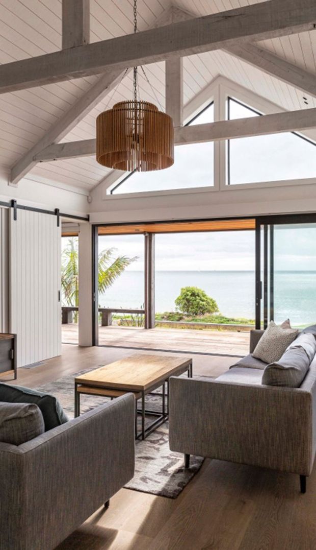 A bright coastal living room with a dramatic pitched, white-lined ceiling and exposed timber structural beams. Large black-framed folding or sliding glass doors open onto a timber deck with an ocean view. The room contains contemporary grey upholstered armchairs and a sofa, a timber coffee table, a patterned rug, and a large woven pendant light.