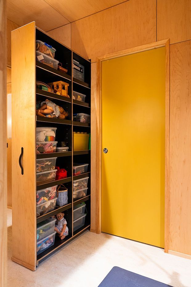 A compact storage area with extensive built-in open shelving and a tall timber cabinet filled with clear plastic storage bins and toys. The walls, ceiling, cabinetry, and door are clad in warm timber, while the adjacent door is a vivid mustard yellow. A small section of blue mat is visible on the pale floor.