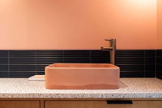 Close-up of a contemporary bathroom vanity featuring a rectangular terracotta-colored vessel basin, brushed metal mixer tap, black horizontal wall tiles, a terrazzo-style benchtop, and timber cabinetry below. The warm peach wall creates a bold, tonal palette.