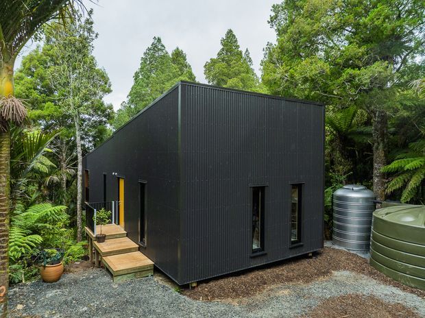 A compact single-storey black corrugated-clad building set within dense native bush and tree ferns. The simple rectangular form has a steep gable roof, narrow dark-framed windows, a small timber entry stair, gravel surroundings, and prominent cylindrical rainwater tanks beside the building.