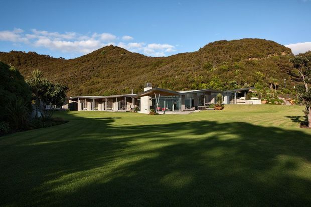 Wide exterior view of a low-profile modern house set against densely vegetated hills, with extensive floor-to-ceiling glazing, flat and gently sloping roof forms, and a broad manicured lawn in the foreground. The architecture is integrated into a natural, wooded landscape.