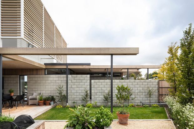 Contemporary multi-level building viewed from a landscaped courtyard, featuring horizontal slatted facade screening, exposed concrete slab rooflines, slender dark metal posts, and pale concrete block privacy walls. The courtyard includes a paved patio, lawn, raised garden beds, potted plants, outdoor seating, and timber fencing.
