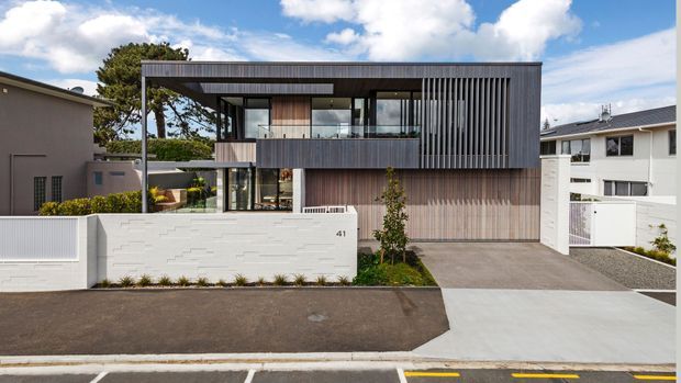 Street-facing exterior photograph of a contemporary two-storey residence with a boxy rectilinear form, dark vertical cladding, timber-toned wall panels, extensive glazing, a projecting upper level, balcony, and an integrated garage. A pale rendered boundary wall and concrete driveway frame the front facade.