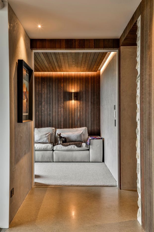 A warm, intimate lounge nook viewed from a polished concrete hallway. The compact room features a light-grey sofa, dark vertical timber wall panelling and matching timber ceiling boards, recessed cove lighting, and a small wall sconce. The surrounding hallway has earthy plastered walls, a low-profile ceiling, and a framed artwork or display cabinet. A cat is seated on the sofa.