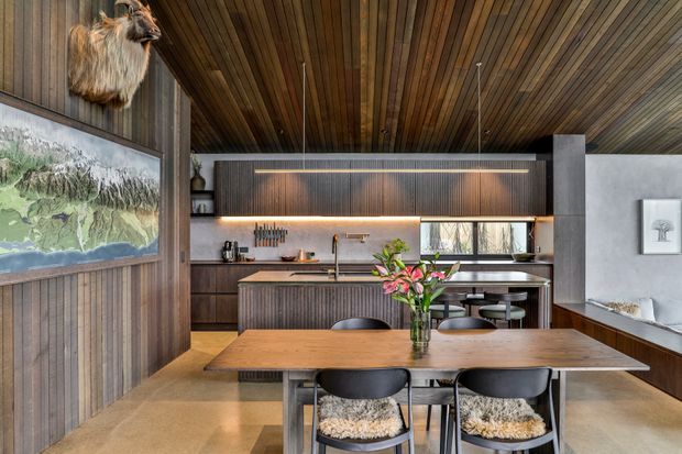 A sophisticated open-plan kitchen and dining area with dark timber wall and ceiling panelling, integrated cabinetry, a large central island, and a timber dining table with black chairs. Warm concealed lighting highlights the ribbed joinery, while a framed landscape artwork and sculptural wall-mounted decor add character. The palette combines rich timber, charcoal, concrete-grey surfaces, and warm neutral flooring.