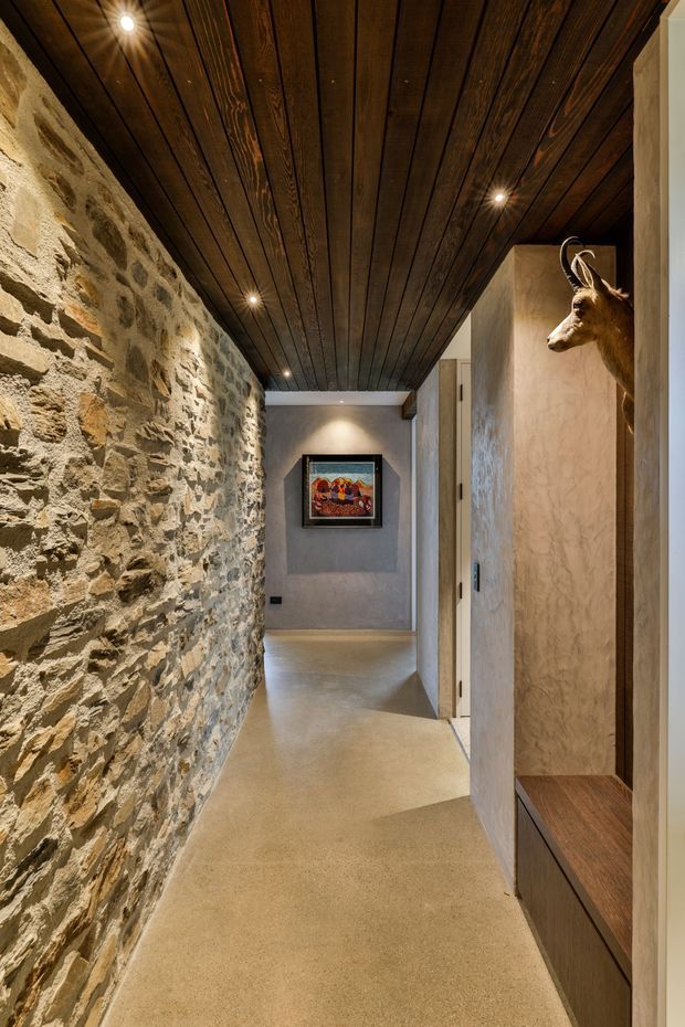 A narrow interior hallway with a rustic exposed stone wall on the left, dark timber-plank ceiling with recessed spotlights, and a warm polished concrete or stone floor. The right side contains built-in dark timber joinery and a textured plaster wall, with a mounted deer head and framed artwork adding decorative detail. The corridor has a warm, earthy, heritage-inspired character with contemporary lighting and finishes.