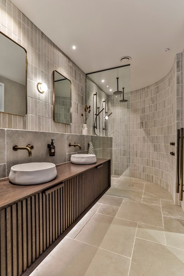 A refined contemporary bathroom with a long double-vanity in dark timber slatted joinery, two white vessel basins, bronze-toned wall-mounted tapware, and rounded rectangular mirrors. The room features large beige stone-look floor tiles, vertically arranged light taupe wall tiles, a curved tiled shower enclosure with dual rain showerheads, warm integrated lighting, and dark metal accents.