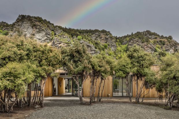 Low, modern building integrated into a rugged mountainous landscape, with weathered timber-clad walls, extensive glazing, and mature sculptural trees screening the facade. A gravel forecourt leads toward the entry, while rocky hills and a rainbow form the background.