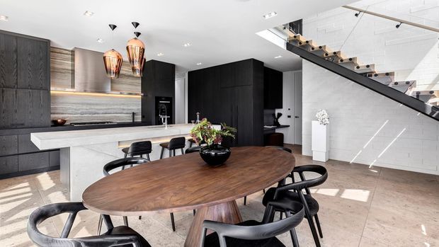 A bright, open-plan contemporary kitchen and dining area with dark timber cabinetry, a long pale stone island, pendant lighting, and a large oval timber dining table with black chairs. A sculptural open-riser staircase with black steel stringer and timber treads rises along a white wall, while daylight enters from large glazing off frame.